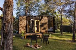 Eine kleine Holzhütte mit einer Tür, einer Bank, zwei Stühlen und einem Feuerkorb im Garten.