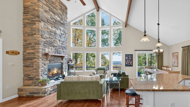 A spacious living room with a stone fireplace, large windows, and a kitchen island. - Mountain retreat w/deck, game room & dog-friendly (Hedgesville)