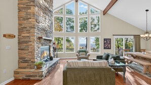 A spacious living room with a stone fireplace, large windows, a ceiling fan, and a dining area. - Mountain retreat w/deck, game room & dog-friendly (Hedgesville)