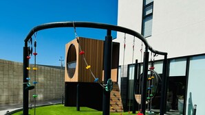 A playground with climbing structures and swings, surrounded by a building with large windows and a grassy area. - Alameda Apartment (Tijuana)
