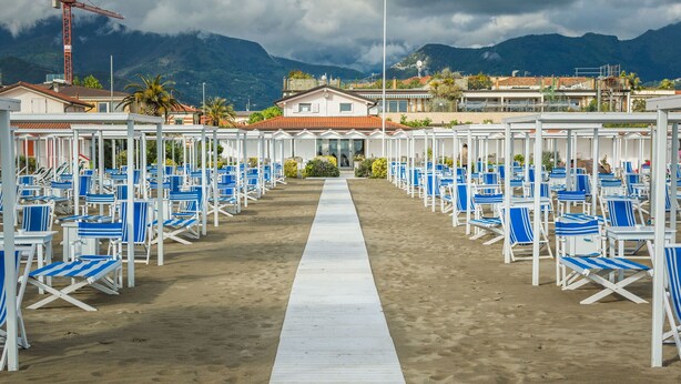 Un hôtel situé en bord de mer, avec des rangées de transats bleus et blancs, une allée centrale et une chaîne de montagnes en arrière-plan.
