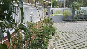 Um pátio pavimentado com plantas em vasos, cerca branca e um edifício com telhado de telhas ao fundo.