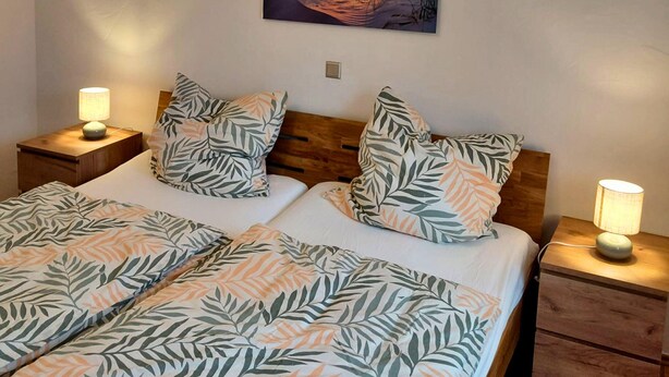 A double bed with leaf-patterned bedding, two bedside lamps, a wooden headboard, and a sunset painting on the wall. - Holiday Home in Birresborn With Balcony (Birresborn)