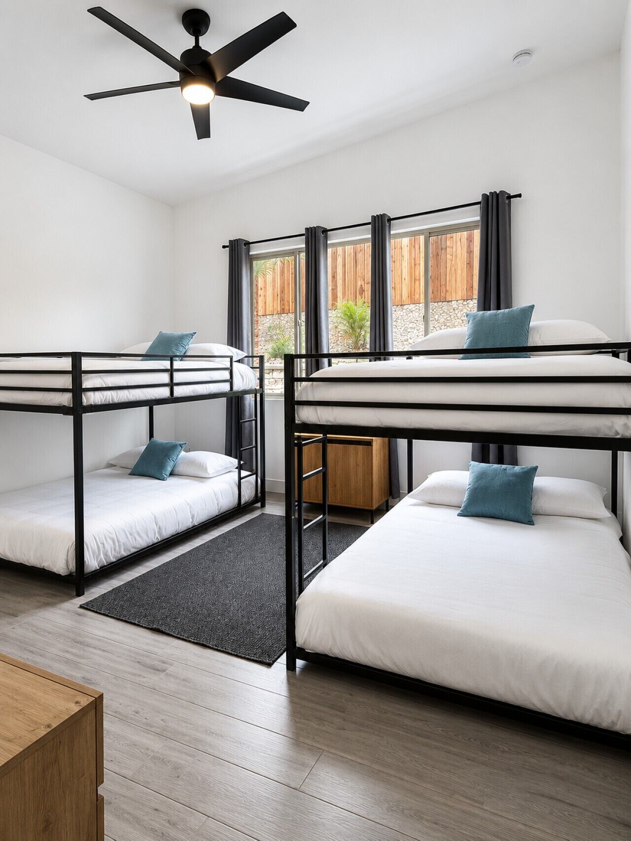 Una habitación con literas, ventilador de techo, ventana con cortinas y suelo de madera.