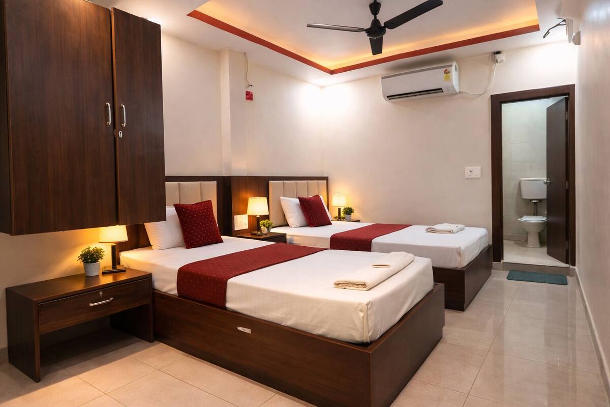 Habitación de hotel con dos camas, una mesita de noche, un escritorio, un ventilador de techo, un equipo de aire acondicionado y un baño visible a través de una puerta abierta.