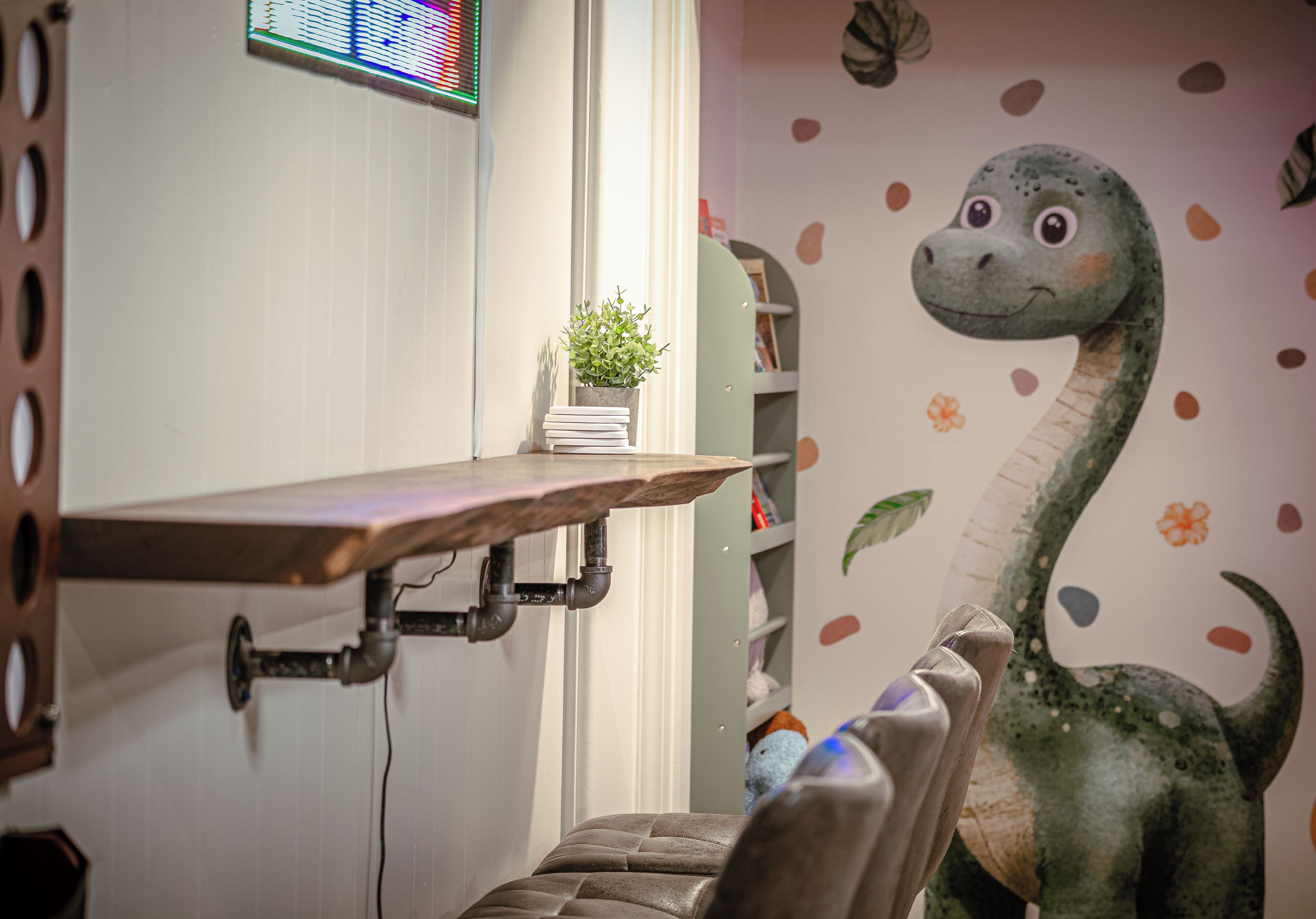 A room with a desk, a potted plant, a dinosaur wall decal, and a bookshelf.