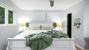 A bedroom with a white bed, two bedside lamps, a green blanket, and a window with blinds. - Grisham Getaway: Hot Tub + Game Room + Firepit + Kid & Pet Friendly (Bella Vista)