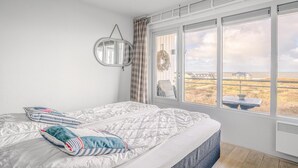 A bedroom with a large window, a bed with striped pillows, a round mirror, and a wreath on the wall. - 4 Person Apartment With sea View (De Koog)