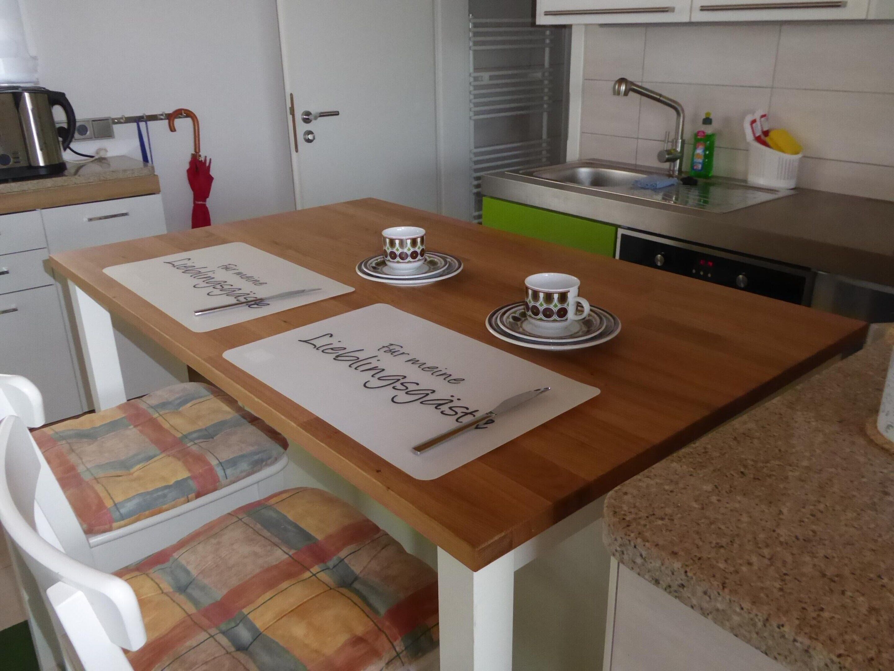 A small kitchen with a wooden table, two placemats, a kettle, a red umbrella, and a sink.
