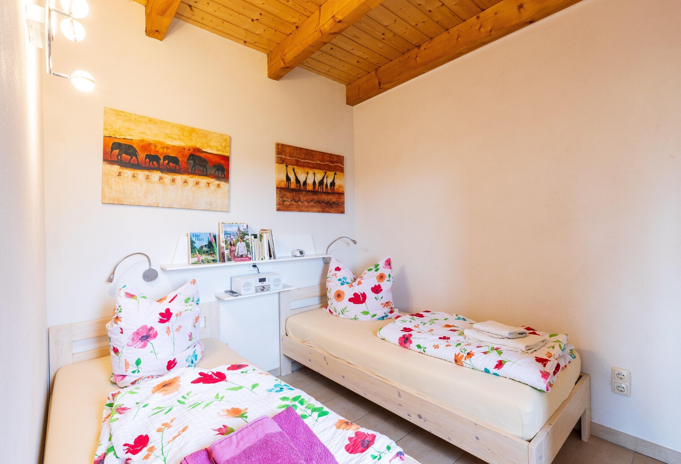 Camera da letto con due letti singoli, testiere in legno, uno scaffale con libri e una piccola pianta, e due opere d'arte a parete raffiguranti elefanti e giraffe.
