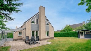Ein zweistöckiges Haus mit überdachter Terrasse, einem kleinen Gartenhaus und einem gepflegten Rasen.