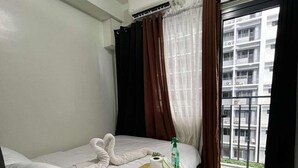 A hotel room with a bed, a bedside table with toiletries, a window with curtains, and a view of a building. - Cozy Haven (Pasay)
