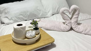 A neatly made bed with white linens, a heart-shaped towel arrangement, a tray with toiletries, and a small plant. - Cozy Haven (Pasay)