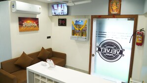 Een hotel receptie met een bruine bank, een wit receptiebalie, een televisie, een klok, een ingelijst schilderij, een decoratieve guirlande en een brandblusser.