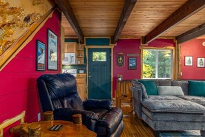 A cozy cabin interior with a wooden ceiling, a red wall, a blue door, a grey sofa, and a black leather chair. - Fir Tree Cabin | Grill, Fireplace (Packwood)