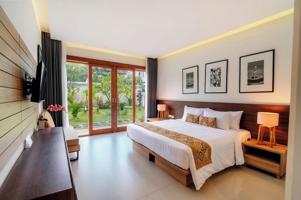 A modern hotel room with a large bed, a desk, a TV, and a sliding glass door to the garden.