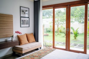 A bedroom with a bed, a sofa, a nightstand, a window with a view of a garden, and framed pictures on the wall. - THE ULU Beach Club and Bungalows (Lembongan Island)