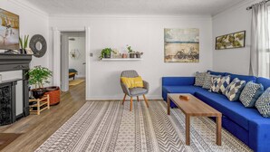 Ein Wohnzimmer mit blauem Sofa, einem Holztisch, einem gemusterten Teppich, einem grauen Sessel mit gelbem Kissen und einem Kamin.