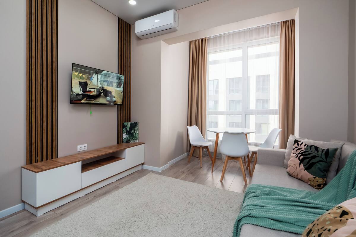 Una sala de estar moderna con un televisor de pantalla plana, una mesa de comedor con sillas, un sofá y un ventanal con cortinas.
