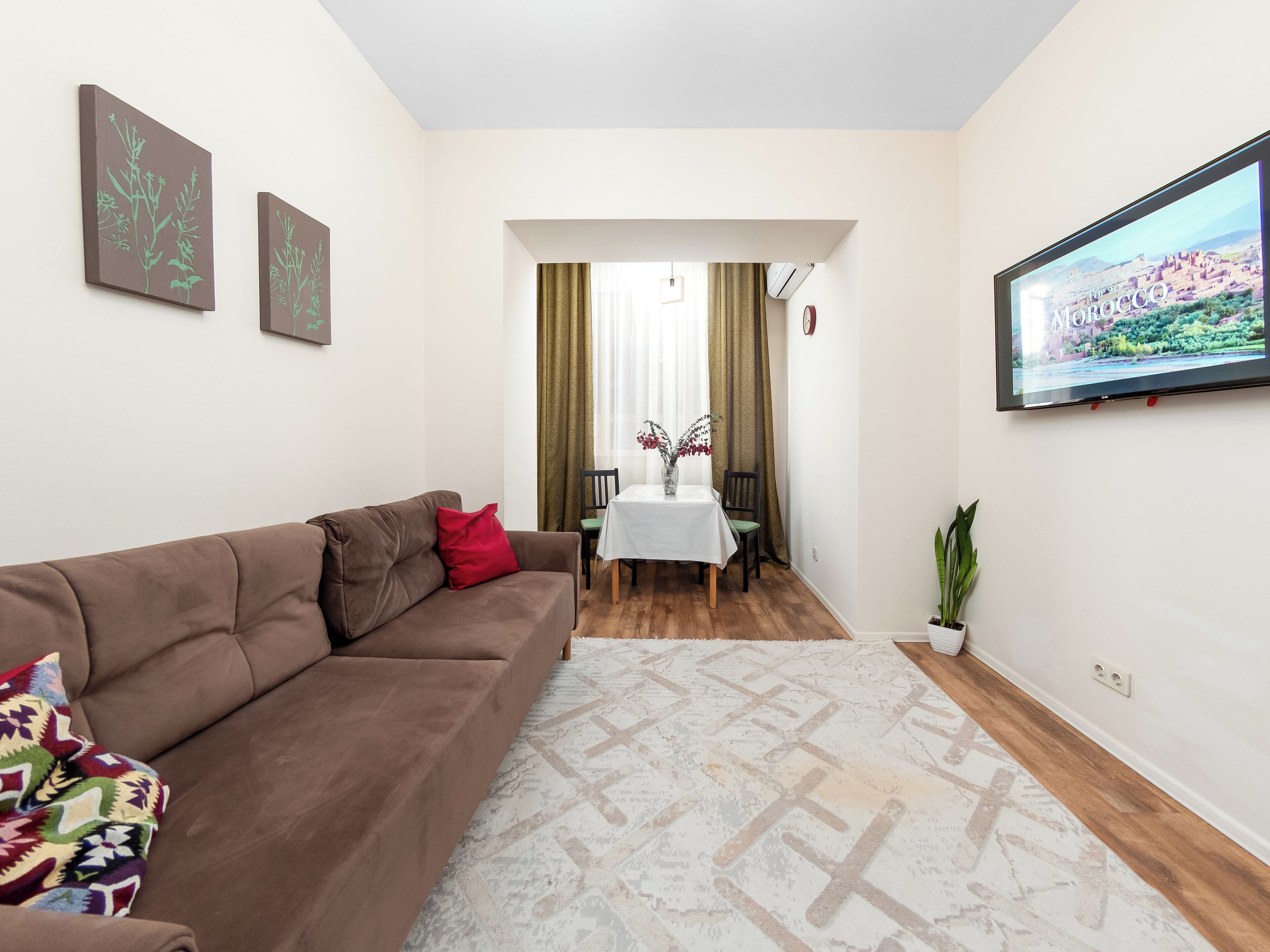 A living room with a brown sofa, a dining table with chairs, a TV on the wall, and a plant.