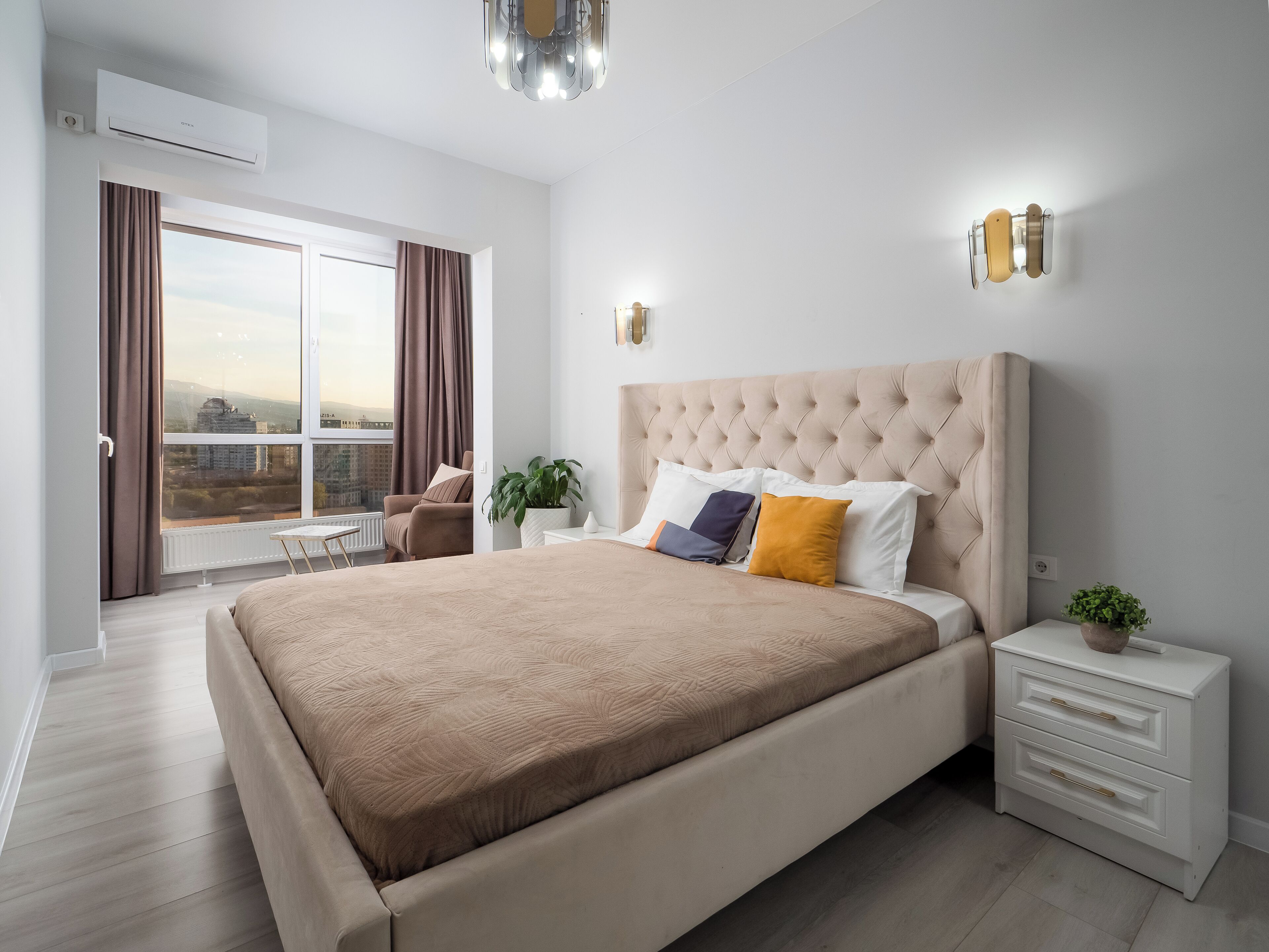 Un dormitorio moderno con una cama grande, una mesita de noche, una planta en maceta y vistas de la ciudad a través de un ventanal amplio.
