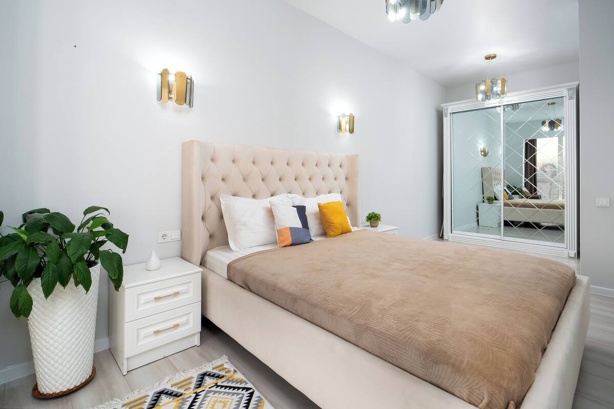 Un dormitorio moderno con cabecera capitonada, una cama grande, mesitas de noche, una planta en maceta y un armario con espejo.