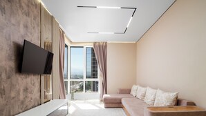 A modern living room with a flat-screen TV mounted on a textured wall, a beige sofa with patterned cushions, and a large window offering a city view. - Business Aparts near MEGA Almaty  (Almaty)