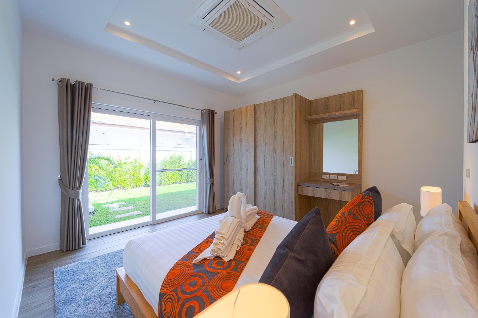 Una habitación de hotel moderna con una cama grande, un armario de madera, un baño con espejo y vistas a un jardín a través de puertas correderas.