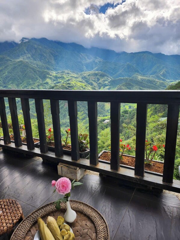 Un balcón con vistas a montañas verdes, una cesta de mimbre con maíz y un jarrón con una flor rosa.