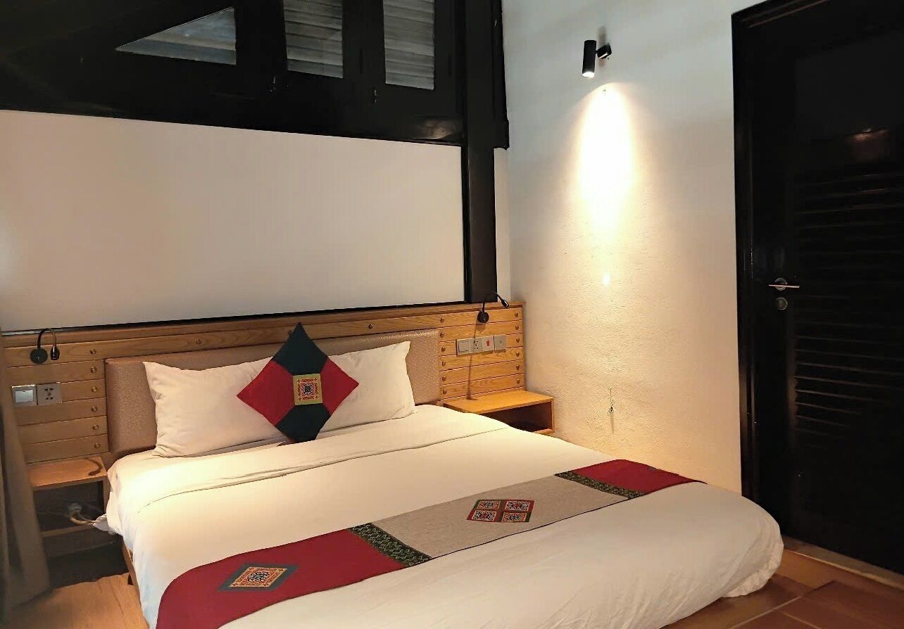 A hotel room with a wooden headboard, a bed with white bedding, a red and black pillow, and a nightstand.