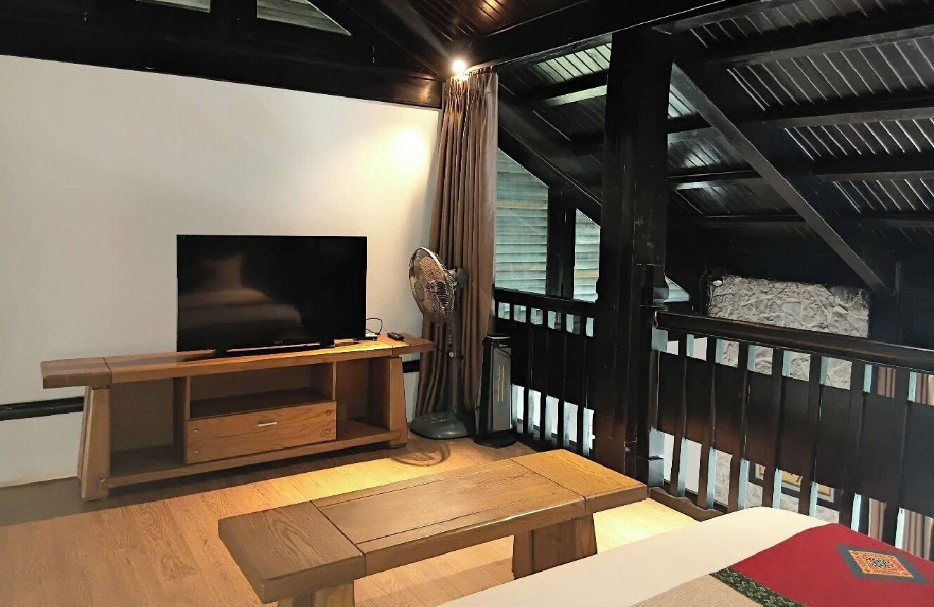 A cozy room with a wooden bench, a TV on a wooden stand, a fan, and a view of a stone wall through a window.
