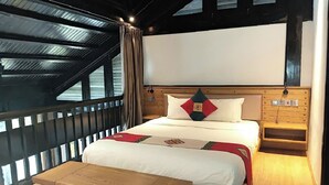 A bedroom with a large bed, a wooden nightstand, a wooden bench, and a view of the outdoors through a window with a metal roof.