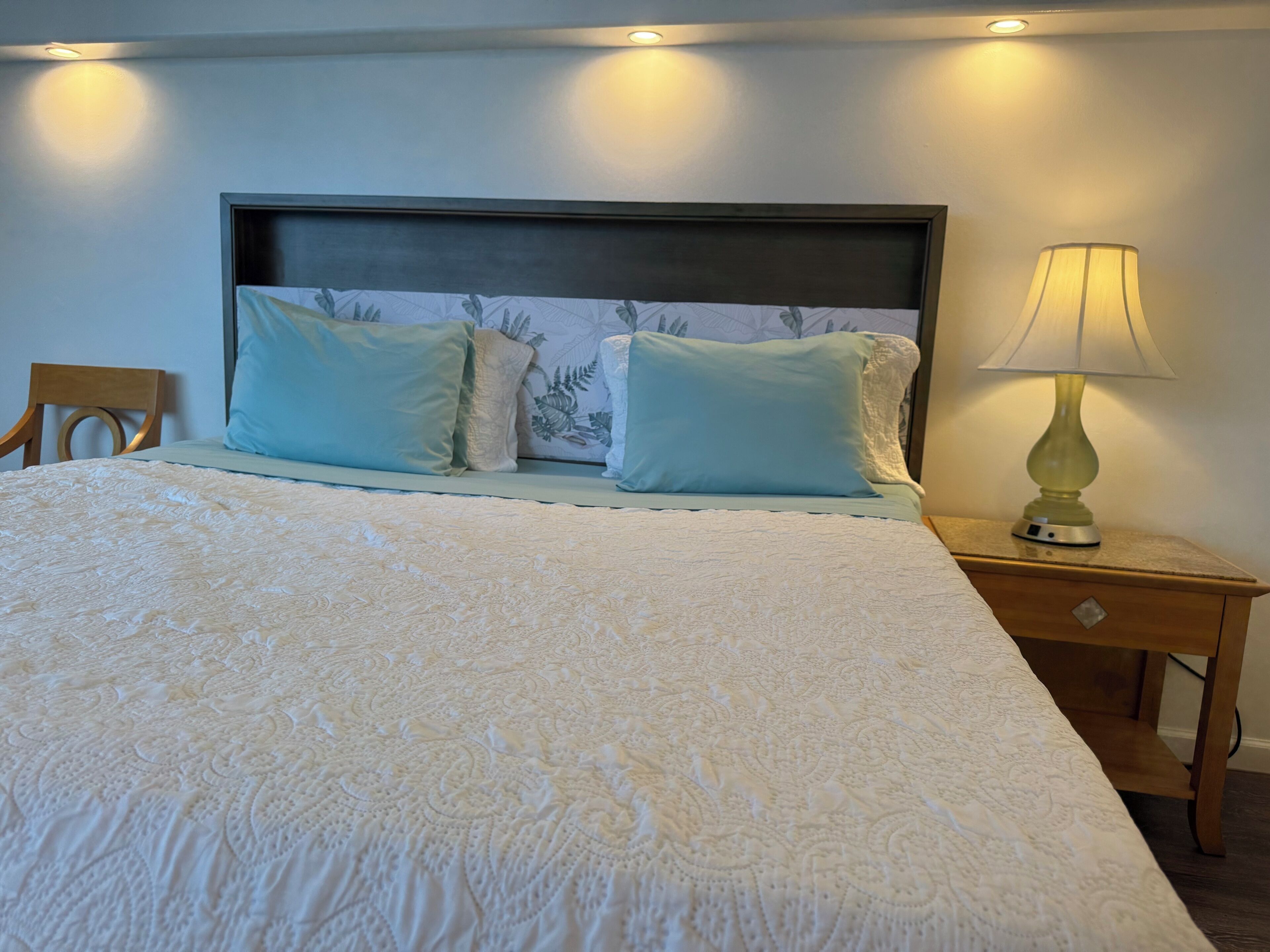 Un lit bien fait, recouvert d’une couvre-lit à motifs, avec deux oreillers, une tête de lit ornée de motifs floraux, une lampe de chevet et une table de chevet en bois.