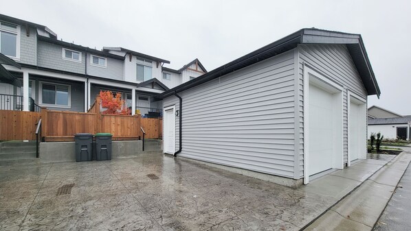 A residential garage with a closed white door, a wooden fence, and two trash bins. - Beautiful Langley Suite by Hwy 1 & Event Centre (Langley Township)