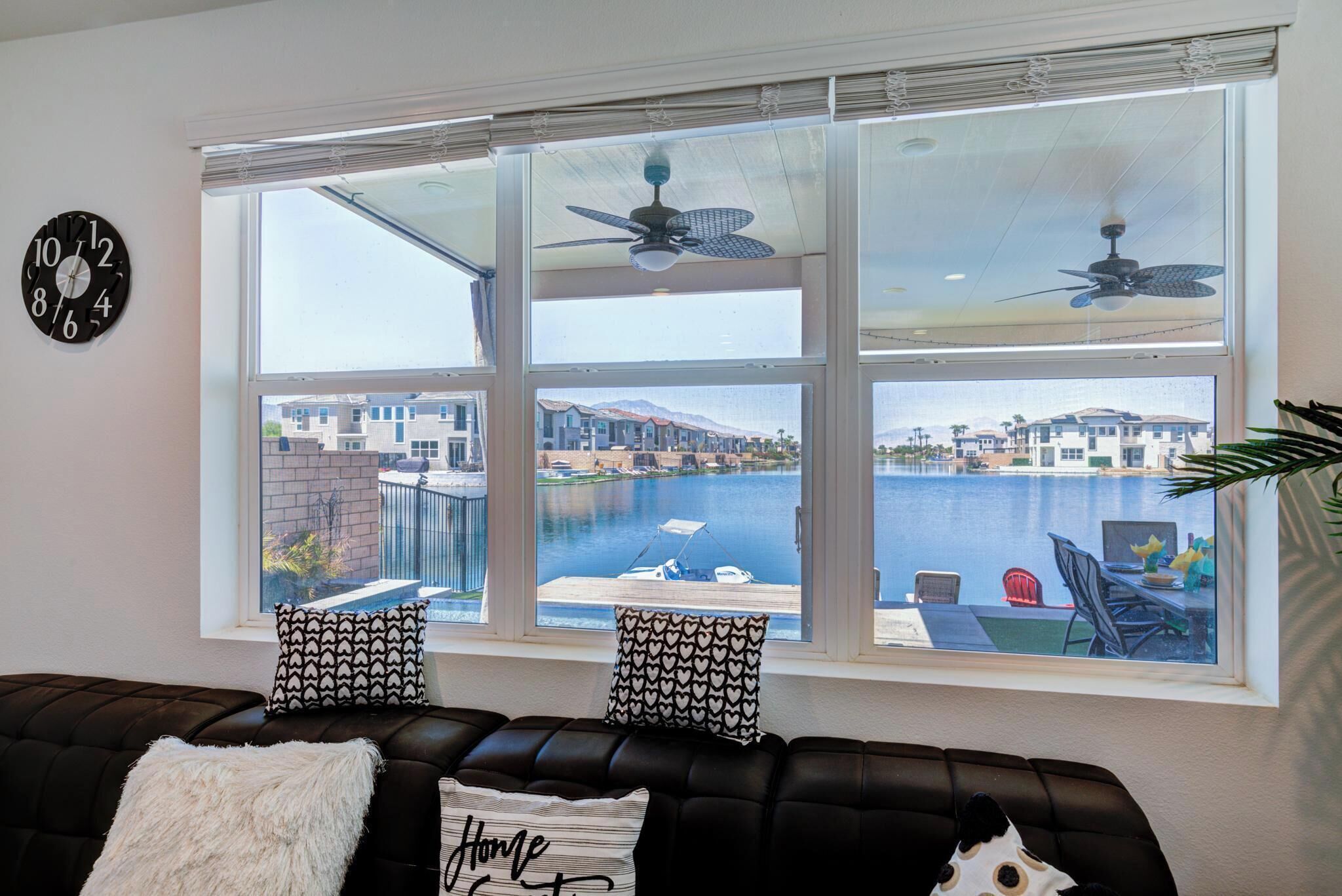 A living room with a large window overlooking a marina and buildings, a black leather sofa, patterned cushions, and a clock on the wall.