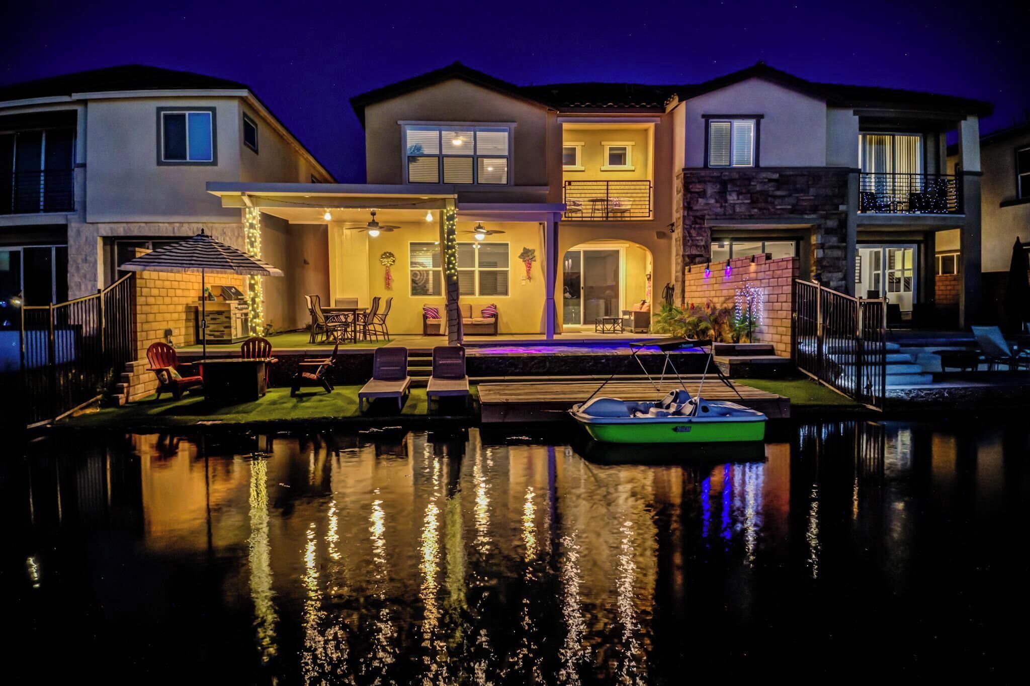 Ein zweistöckiges Haus mit Terrasse, Sitzgelegenheit im Freien und einem Boot, das anleg am Wasser liegt.