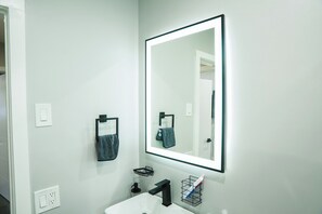 Une salle de bain moderne avec un lavabo blanc, un robinet noir et un grand miroir à cadre noir. On y trouve un porte-serviettes avec une serviette foncée et une petite étagère avec un support à brosse à dents.