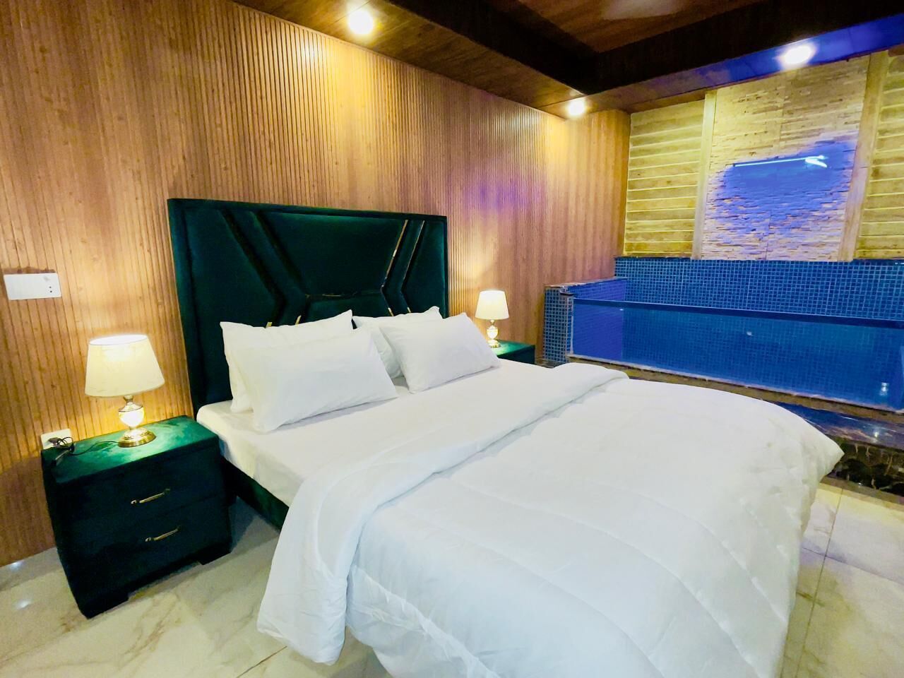 A hotel room with a large bed, wooden panel walls, a nightstand with a lamp, and a view of a pool.