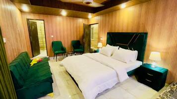 A hotel room with a large bed, a green sofa, two chairs, a nightstand with a lamp, and wooden paneling on the walls.