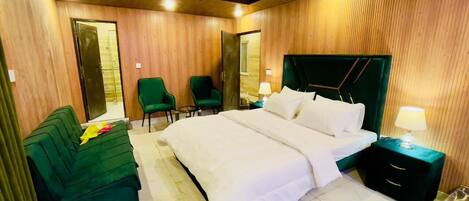 A hotel room with a large bed, a green sofa, two chairs, a nightstand with a lamp, and wooden paneling on the walls.