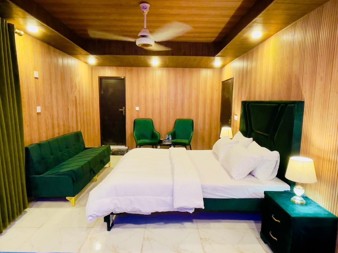Una camera d'albergo con un letto grande, due poltrone verdi, una ventola a soffitto e un comodino con una lampada.