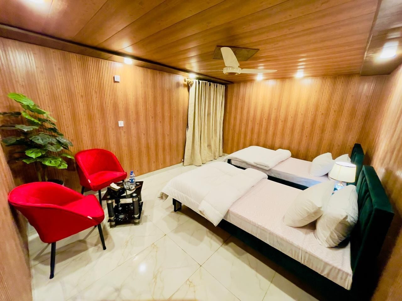 Una habitación con una cama, dos sillas rojas, una mesita de noche con una lámpara y un ventilador de techo.