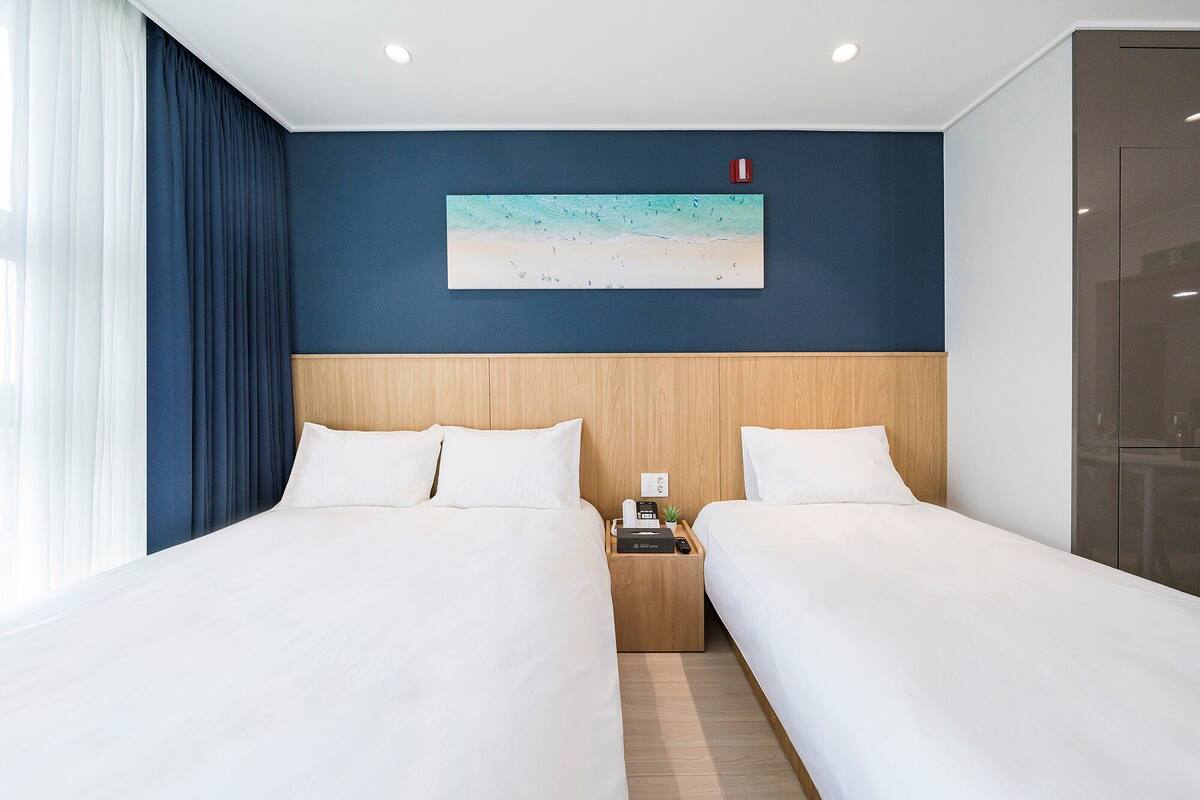 Habitación de hotel con dos camas, cabecera de madera, un cuadro de una playa y una mesita de noche con teléfono y control remoto.