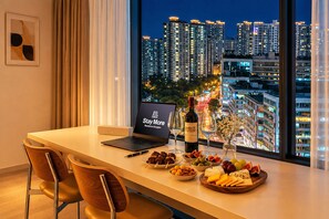 Una habitación de hotel moderna con una computadora portátil mostrando 'Stay More', una botella de vino y una bandeja de frutas sobre una mesa junto a una ventana con vista a la ciudad.