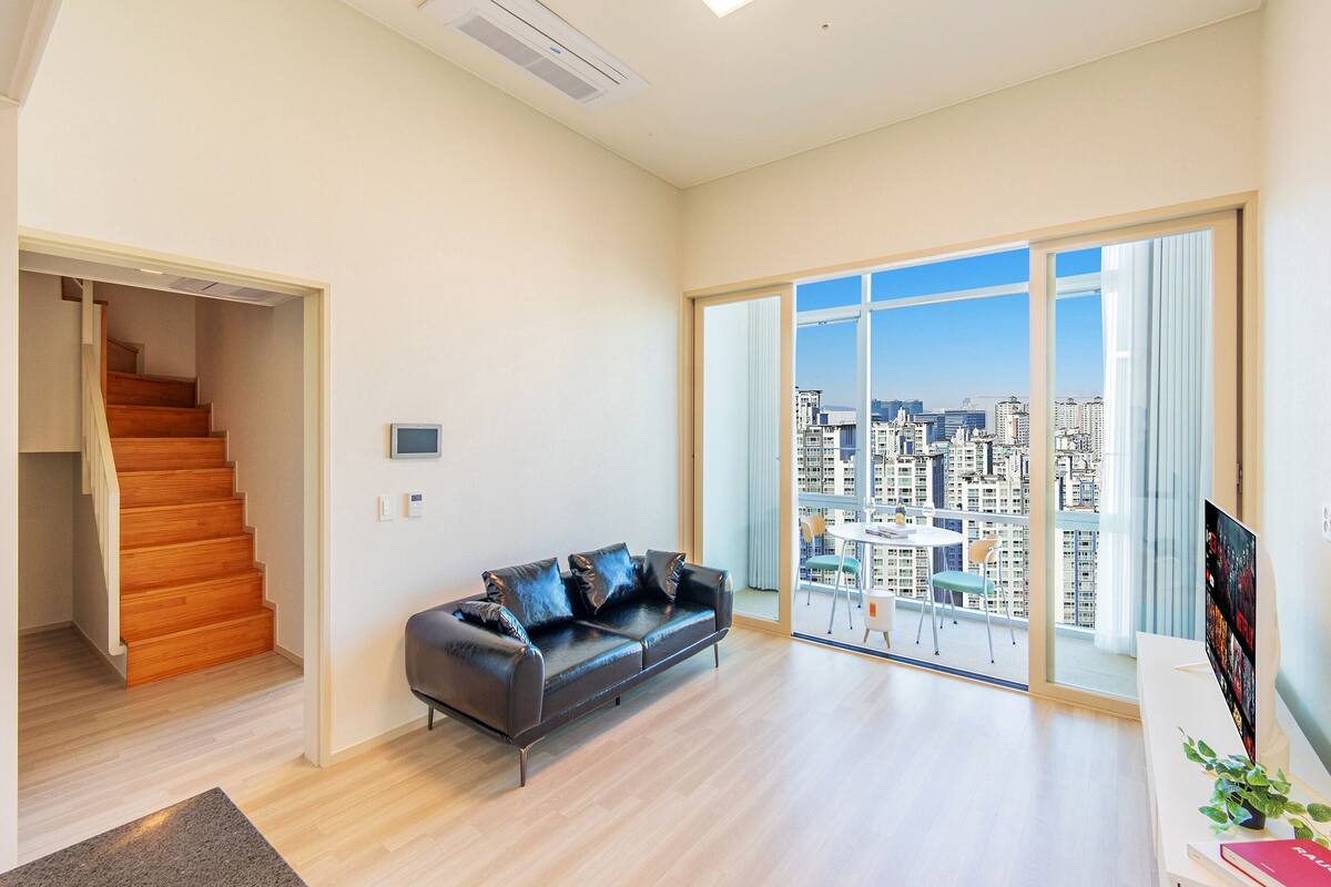 Una sala de estar moderna con un sofá de cuero negro, una escalera y un ventanal con vistas a la ciudad.