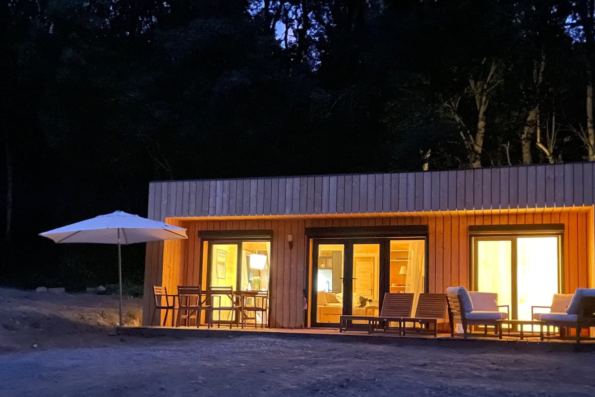 Une cabane moderne dotée de grandes portes vitrées, d’un espace salon extérieur et d’un parasol de patio.