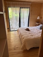Una habitación de madera con una cama, una ventana con cortinas traslúcidas y una lámpara en una mesita.