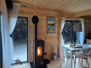 Interior acogedor de una cabaña con estufa de leña, zona de comedor y ventana con vistas a árboles nevados.