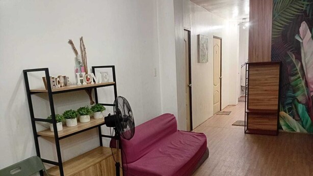 Een kamer met een houten vloer, een rode bank, een zwarte ventilator, een groene stoel en een houten kast.