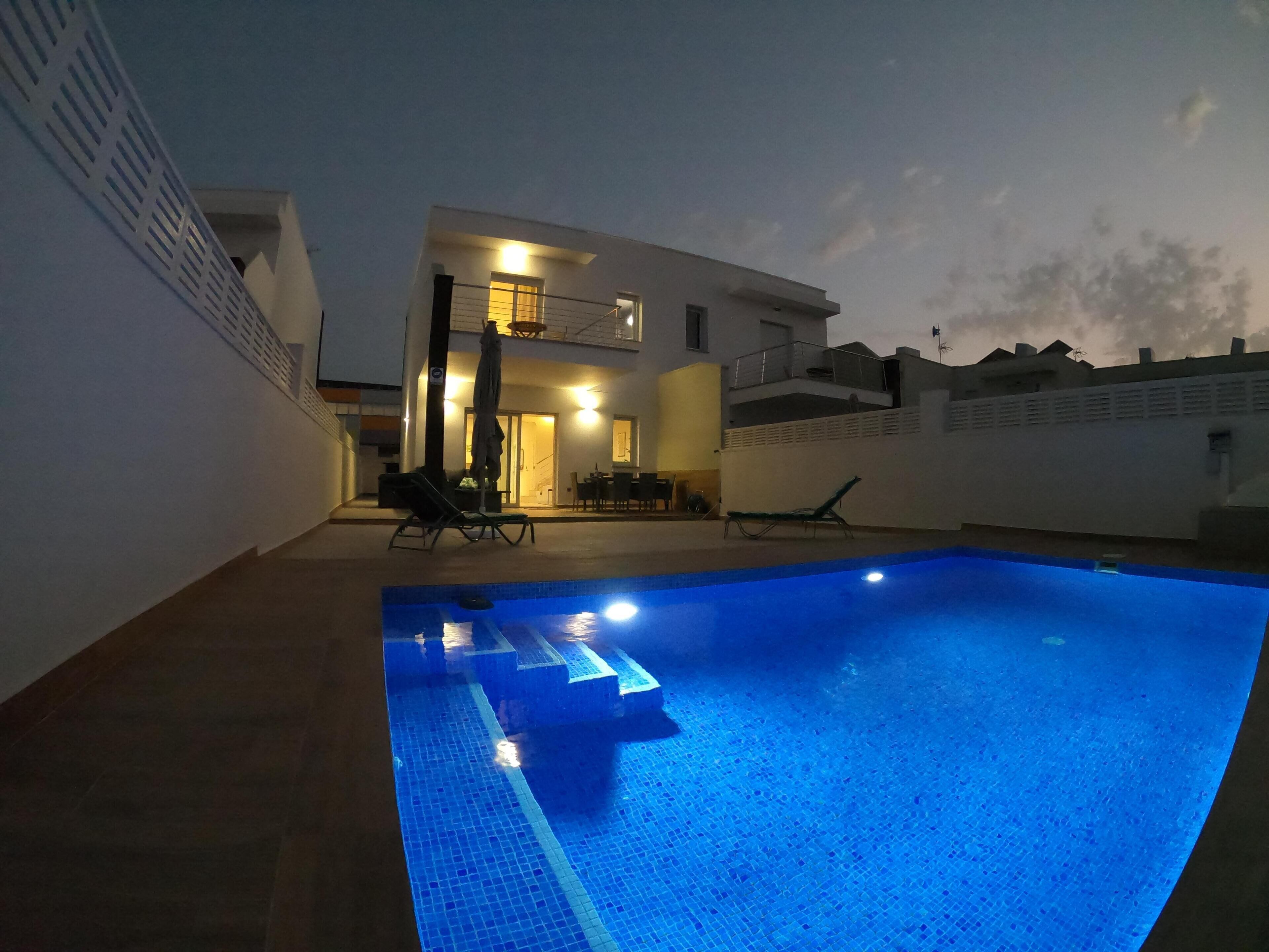Ein Schwimmbecken mit Fliesenbord, ein überdachter Sitzbereich mit Stühlen und einem Sonnenschirm sowie ein zweistöckiges Gebäude mit Balkon im Hintergrund.